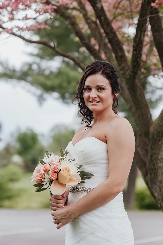 Bride with long dark curls holds a bouquet of white and pink flowers in front of a tree
